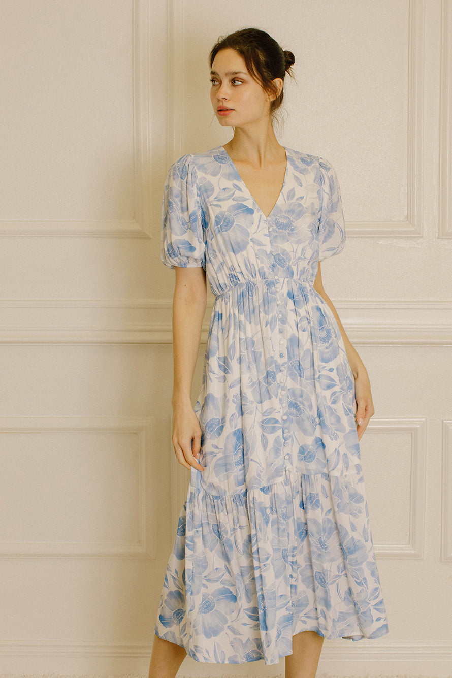 A woman wearing a light blue and white floral midi dress with puff sleeves and a V-neckline, standing against a cream-colored paneled wall.