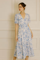A woman wearing a light blue and white floral midi dress with puff sleeves and a V-neckline, standing against a cream-colored paneled wall.