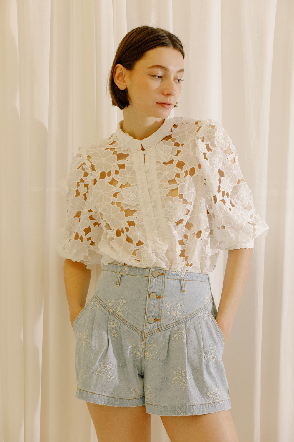 A woman wearing a white floral eyelet short-sleeve blouse and light blue denim shorts with embroidered details, standing in front of a soft cream curtain backdrop.
