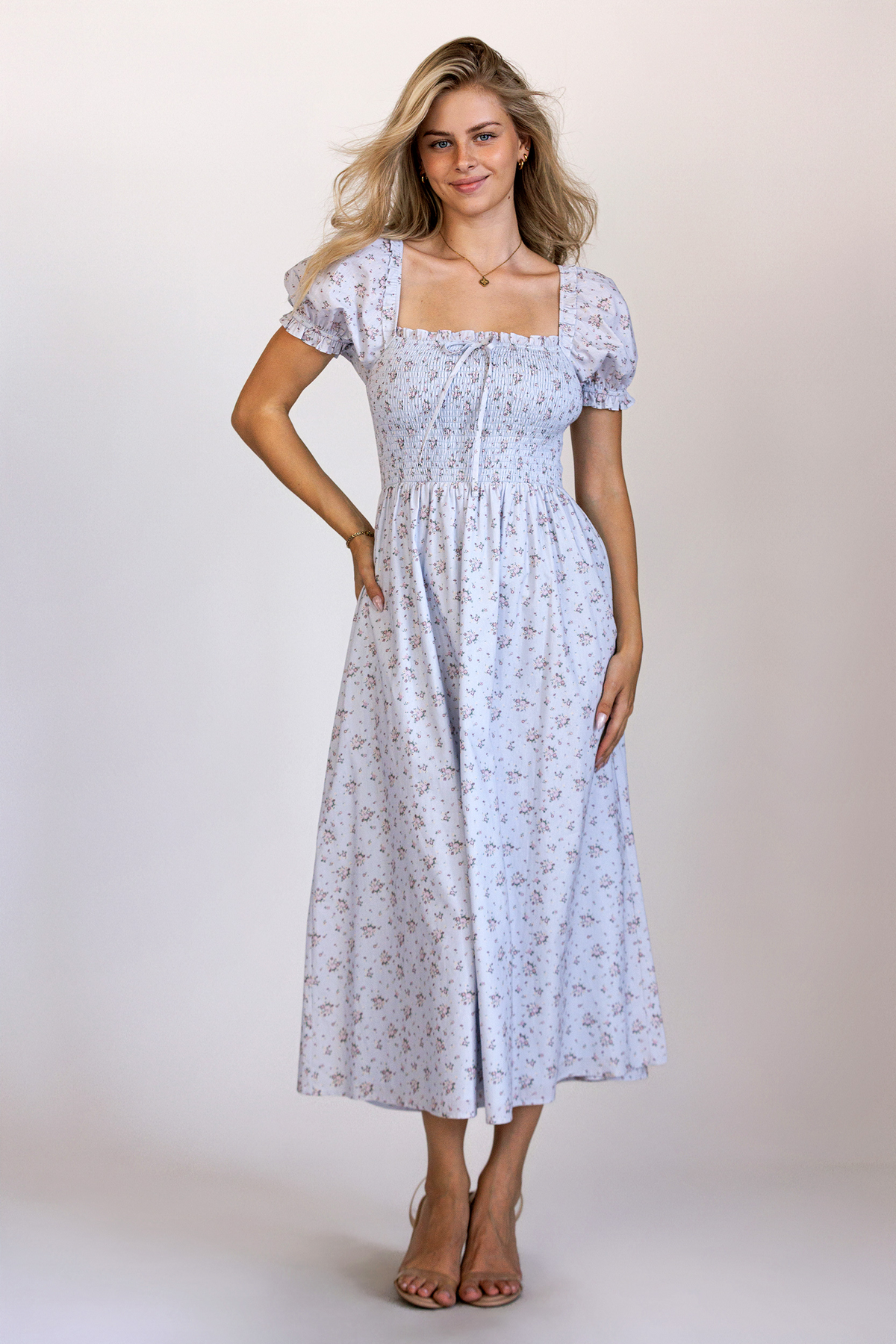 A model wearing a charming blue base midi dress with pink florals, a fully smocked bodice, puff sleeves, and a flowy midi skirt.