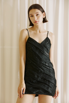 A woman modeling a black spaghetti-strap mini dress with a ruched, draped design and subtle sequin details, set against a soft beige curtain background.