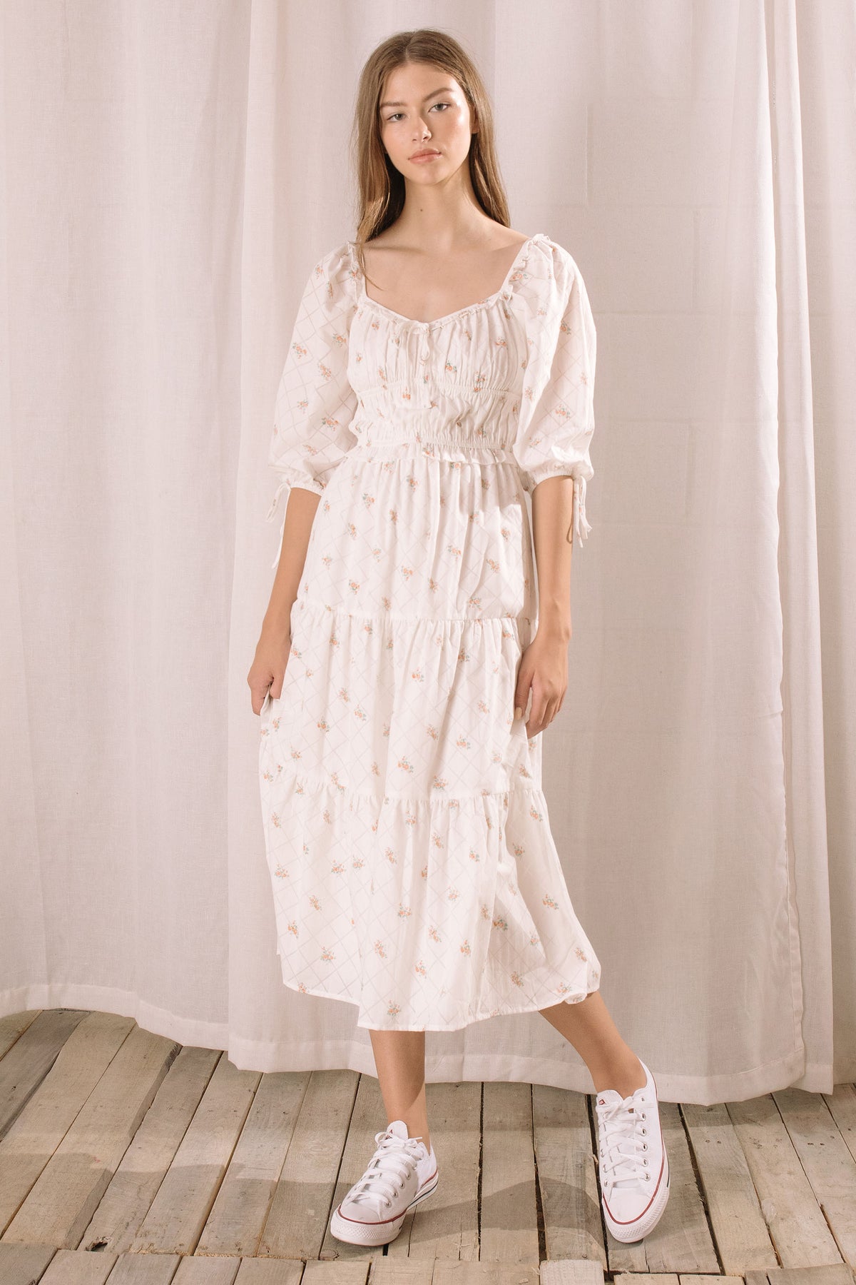 Woman wearing a white dress with a floral pattern and puffed sleeves against a white curtain background