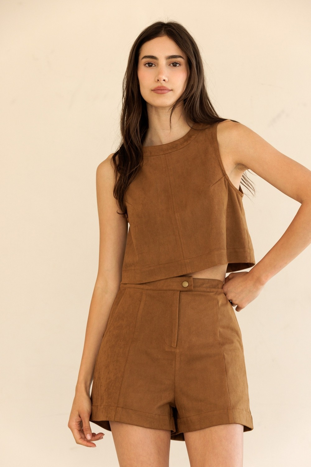 A woman modeling a matching brown faux-suede two-piece set consisting of a sleeveless crop top and high-waisted shorts, set against a plain beige background.
