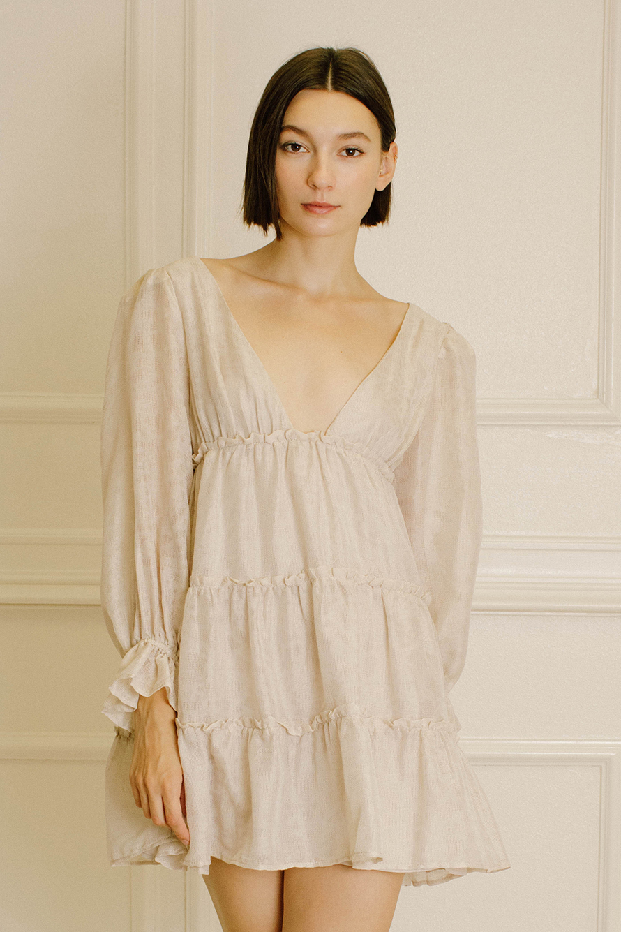 A woman with short dark hair wearing a light beige tiered mini dress with long sleeves, standing against a cream-colored paneled wall.