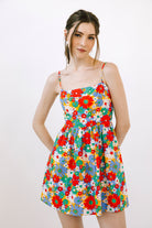 A woman modeling a sleeveless spaghetti-strap mini dress with a vibrant multicolor floral print on a white background, photographed against a clean white studio backdrop.