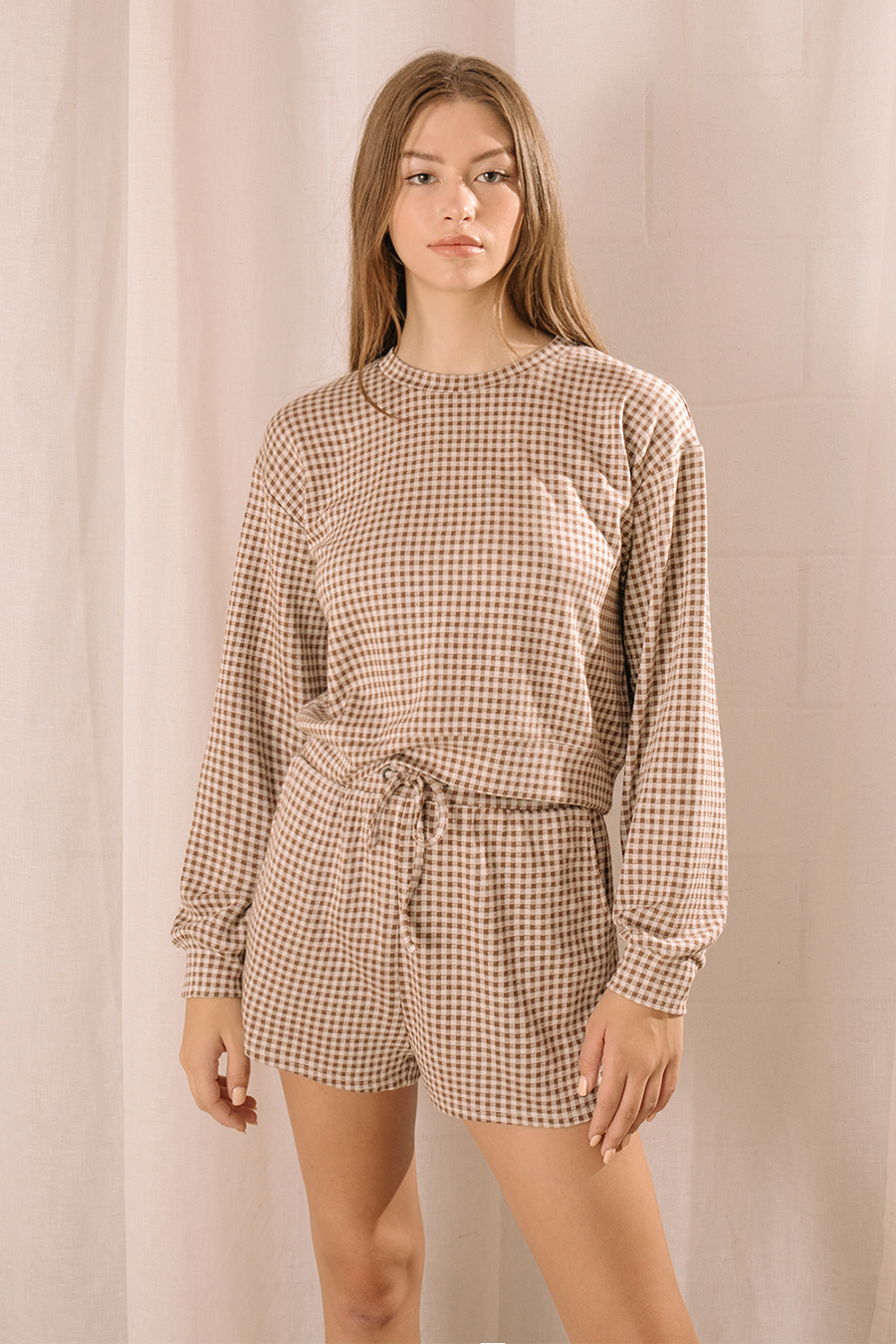 A woman modeling a lounge set that features a relaxed long-sleeve top and high-waisted drawstring shorts in a neutral brown and ivory gingham print.