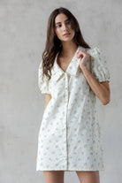A woman modeling a short puff-sleeve button-front dress with a pointed collar and a small blue floral print on a warm off-white background, set against a textured light gray wall.