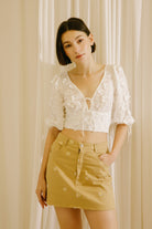 A woman modeling a white lace crop top with dimensional flower embellishment, puff sleeves and a tan mini skirt with white embroidered details, standing in front of a cream-colored curtain backdrop.
