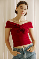 A woman wearing a burgundy off-shoulder rosette top with a round neckline and a central rose detail.