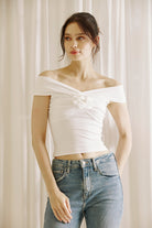 Woman wearing a white off-shoulder top and blue jeans against a light curtain background