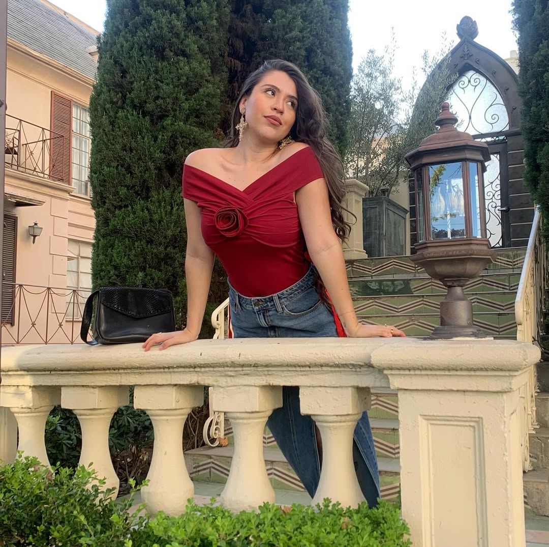 Woman in a burgundy top and blue denim skirt standing on a stone ledge with greenery and a building in the background.