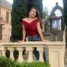 Woman in a burgundy top and blue denim skirt standing on a stone ledge with greenery and a building in the background.