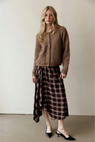 A model wearing a plaid midi skirt that features a brown and beige print, bias-cut silhouette, and a flowy asymmetrical handkerchief hem.