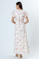 Back view of woman wearing a pink and white floral dress with flutter sleeves on a white background