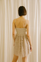 Back of a woman wearing a beige dress with a ruffled skirt against a white curtain background