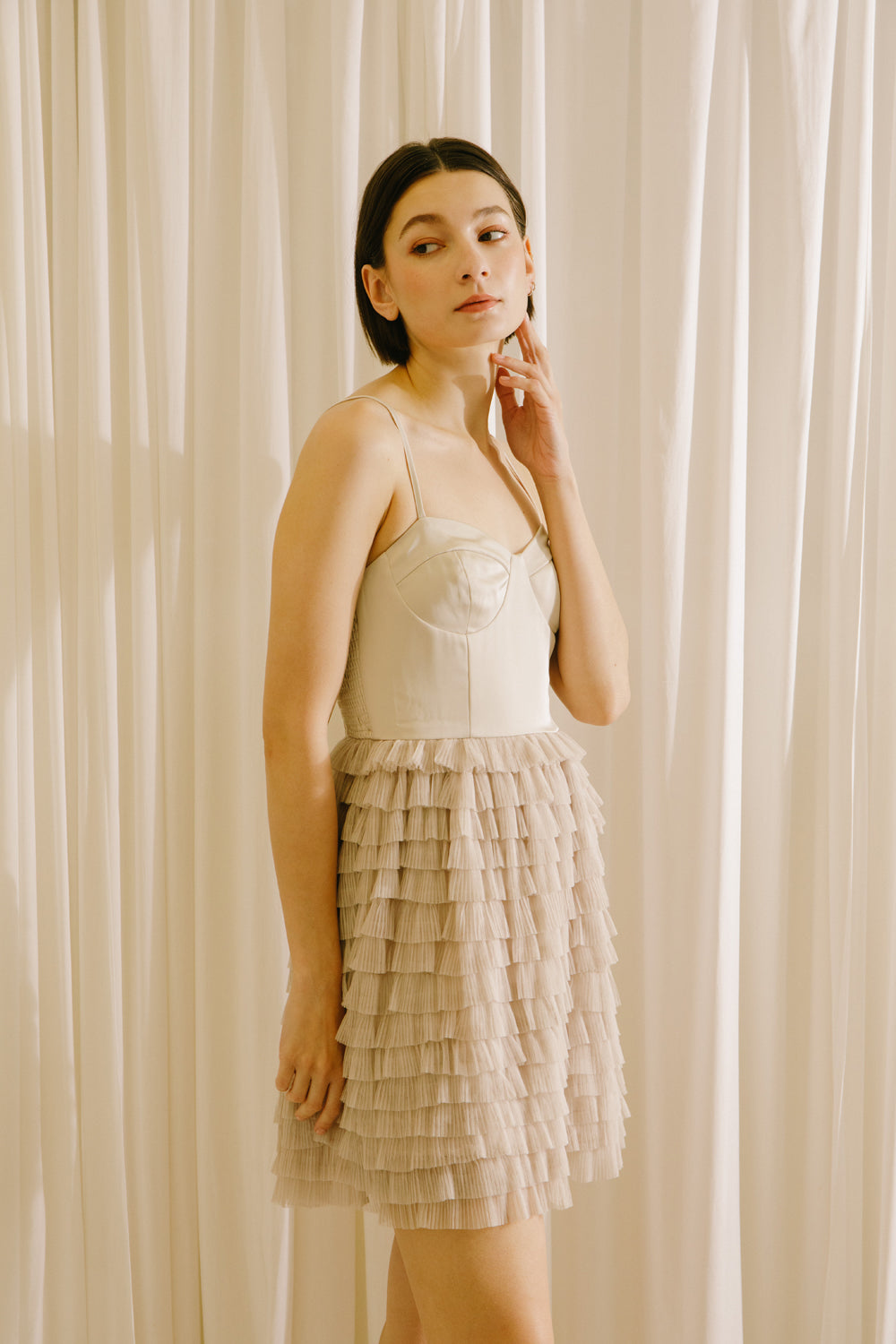 Side of a woman wearing a beige dress with a ruffled skirt against a white curtain background