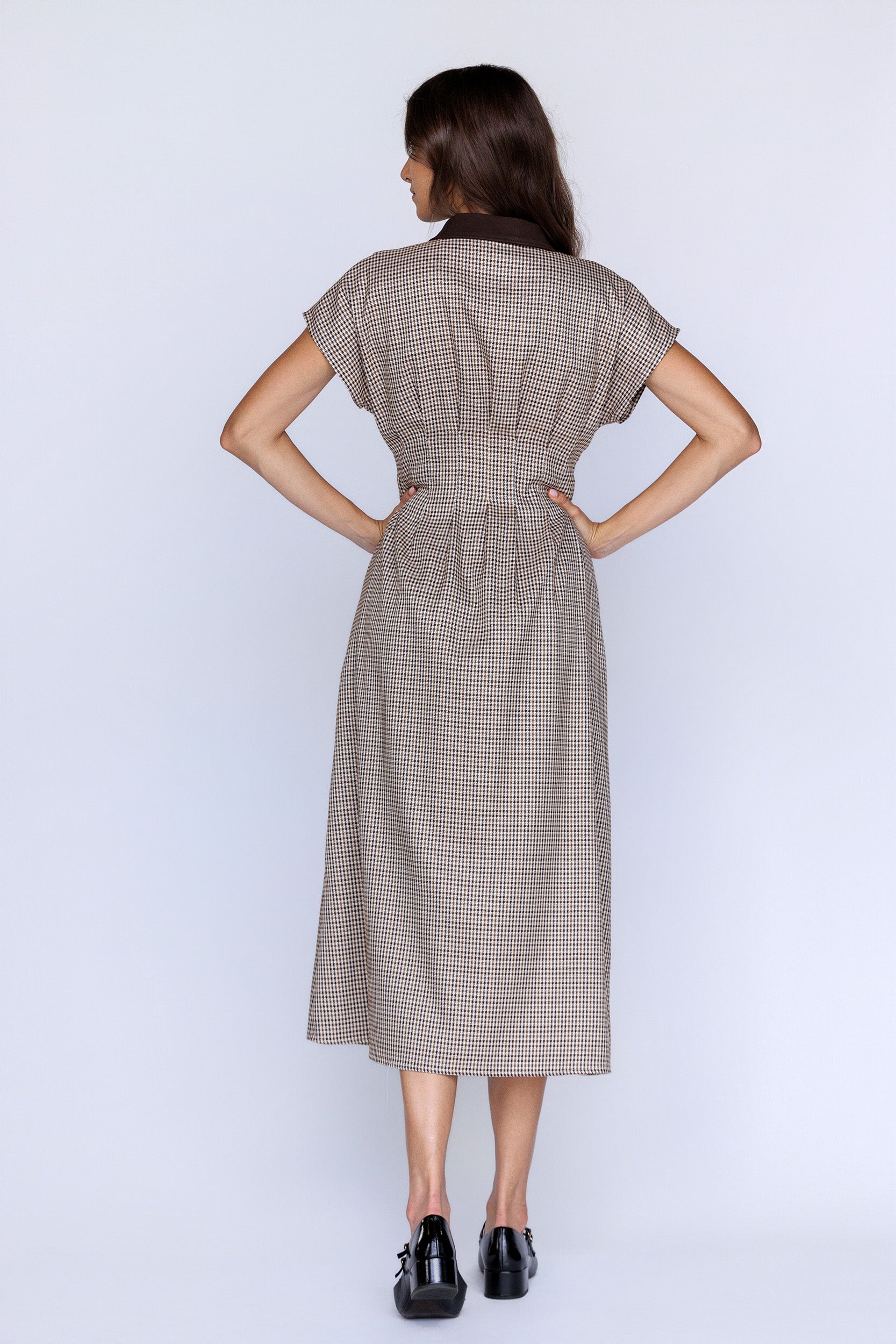 Back view of a woman wearing a brown plaid dress with her hands on her hips against a white background