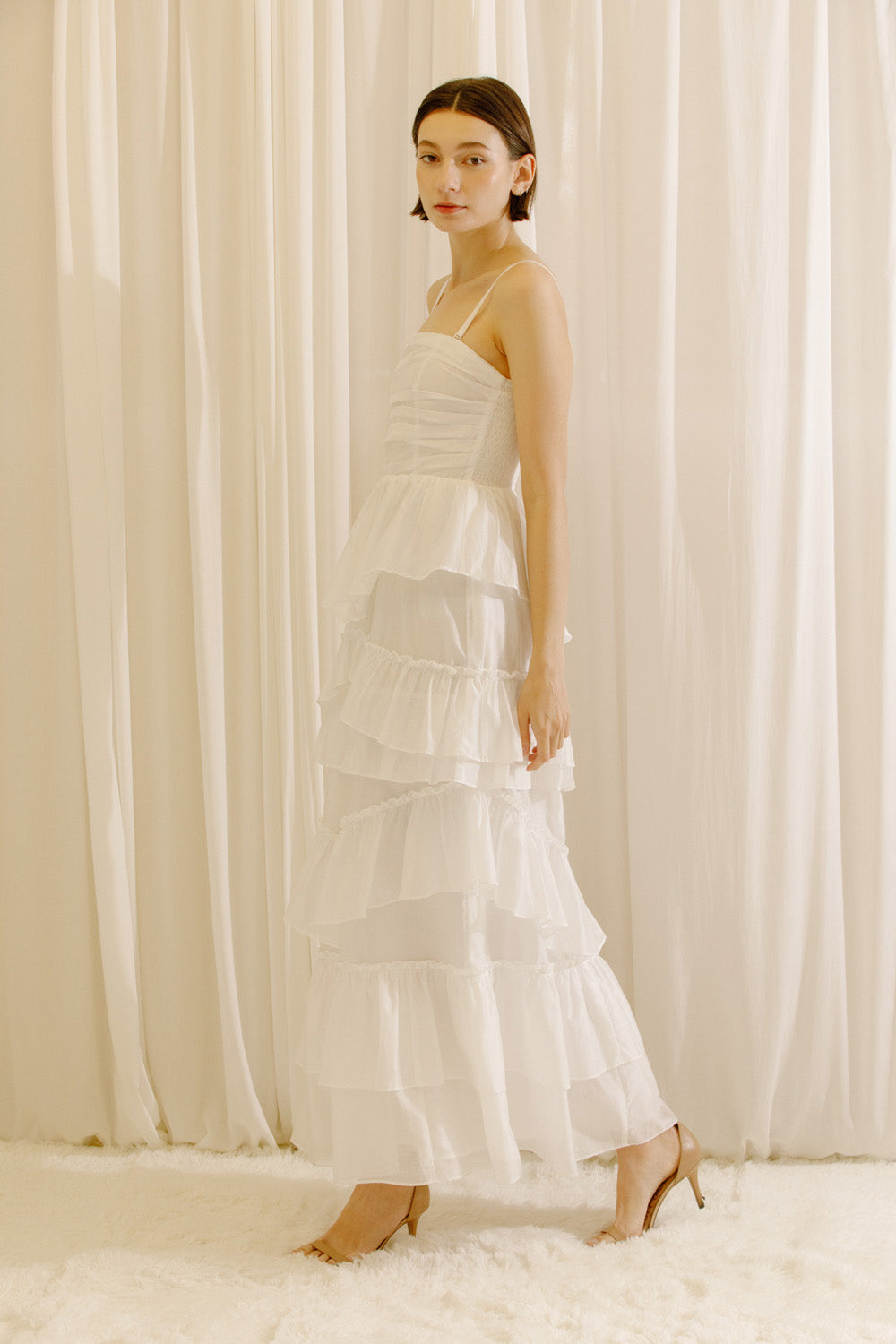 Side view of a woman wearing a white maxi dress standing against a beige curtain backdrop