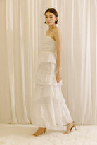 Side view of a woman wearing a white maxi dress standing against a beige curtain backdrop