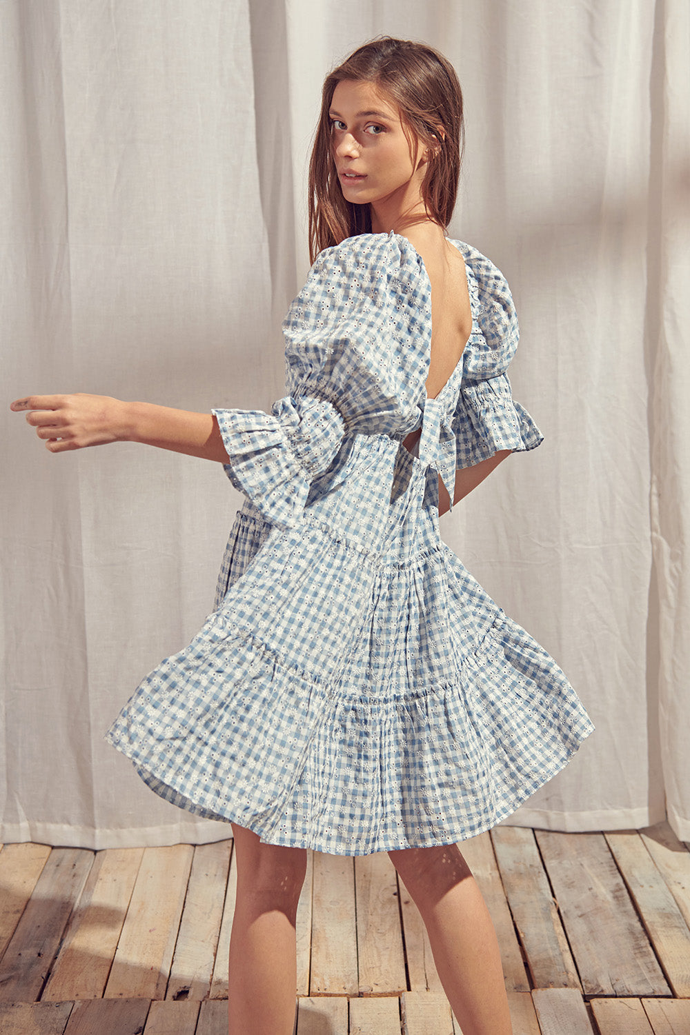 Woman wearing a blue and white checkered dress with a backless design, standing on a wooden floor with a light curtain background.
