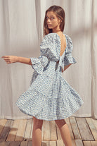 Woman wearing a blue and white checkered dress with a backless design, standing on a wooden floor with a light curtain background.