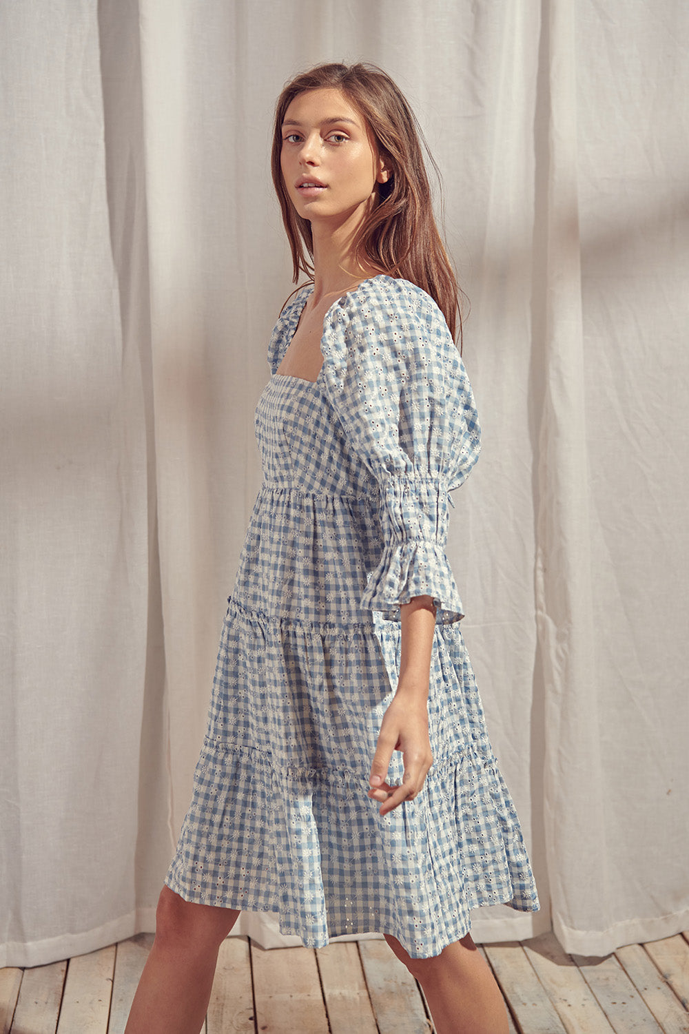 Side view of a woman wearing a blue and white checkered dress with a backless design, standing on a wooden floor with a light curtain background.