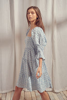 Side view of a woman wearing a blue and white checkered dress with a backless design, standing on a wooden floor with a light curtain background.