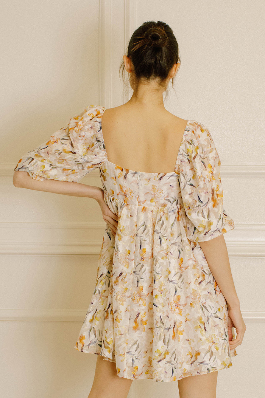 Back view of a woman wearing a beige A-line silhouette mini dress with brown and orange floral pattern dress standing against a neutral wall