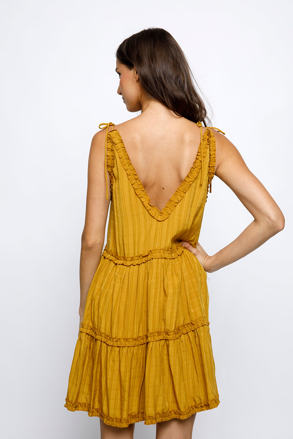 Back view of a woman wearing a mustard yellow dress with ruffled details on a white background