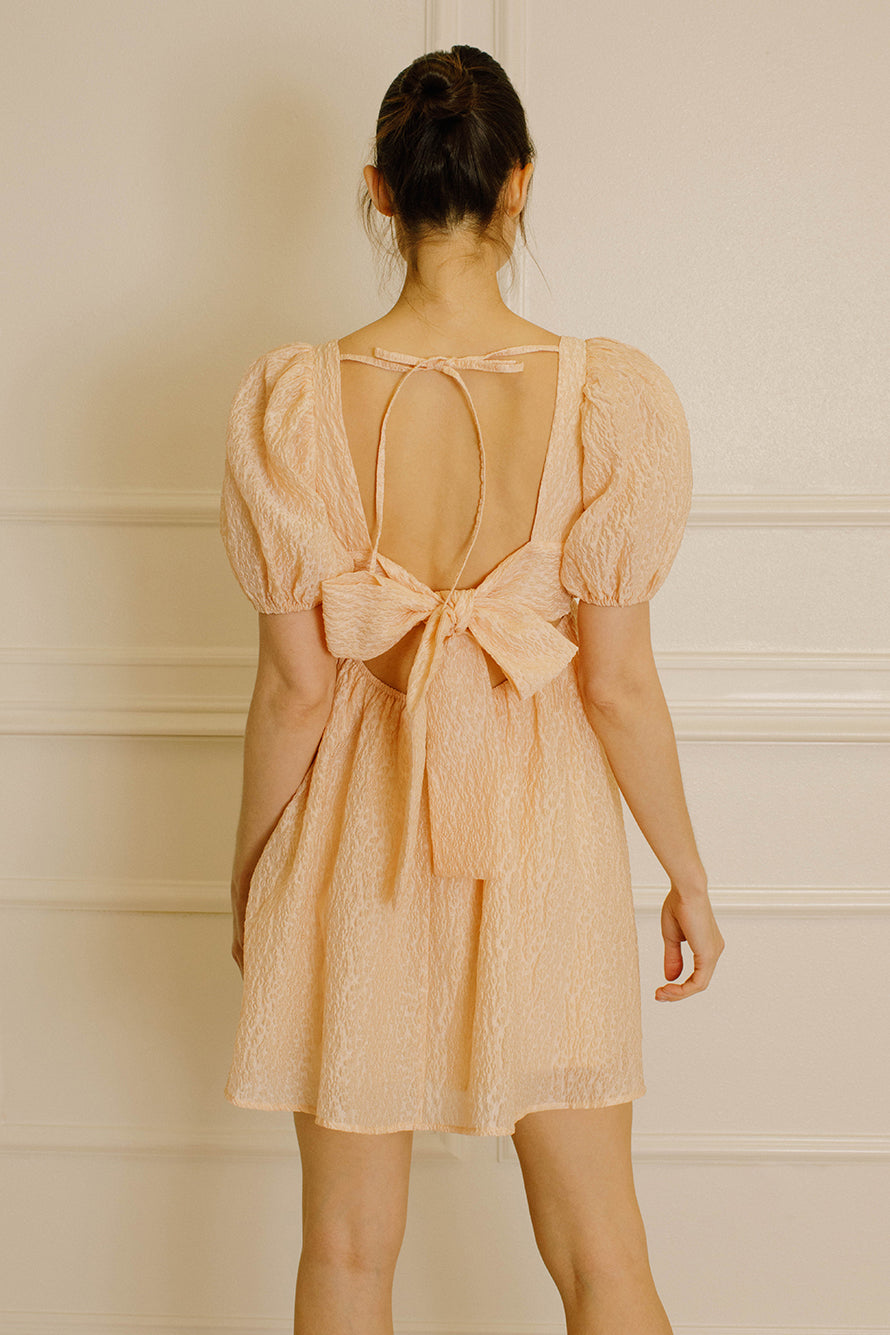 Woman wearing a peach-colored dress with an open back with two self-tie bows and beige high-heels standing against a white paneled wall