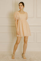 Woman wearing a peach-colored dress and beige high-heels standing against a white paneled wall.