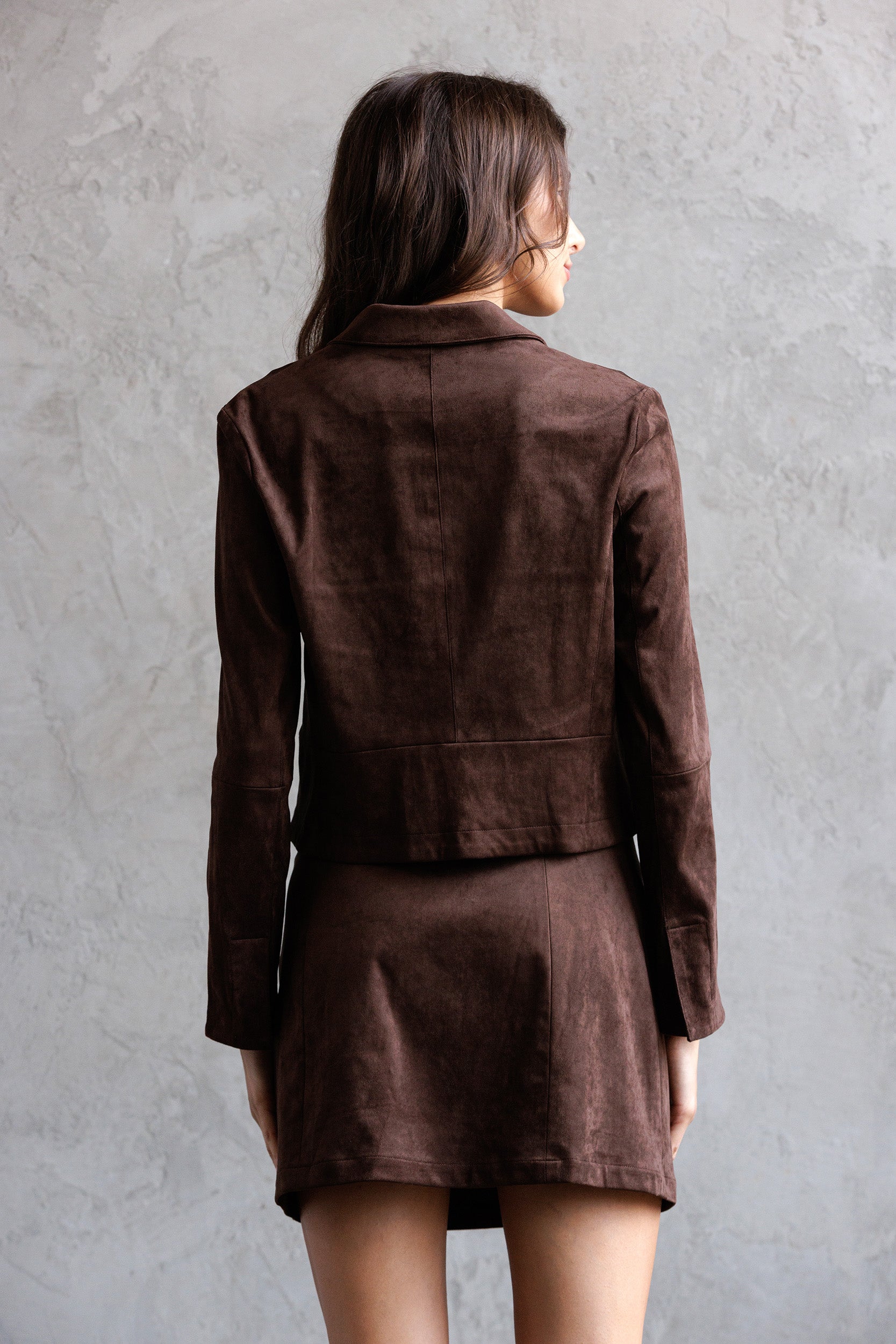Back view of a woman wearing a brown jacket and skirt set against a gray background