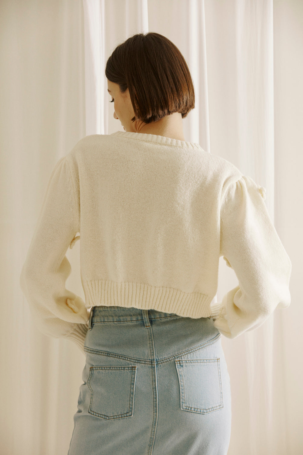 Back view of a woman wearing a cream-colored cardigan against a white curtain background