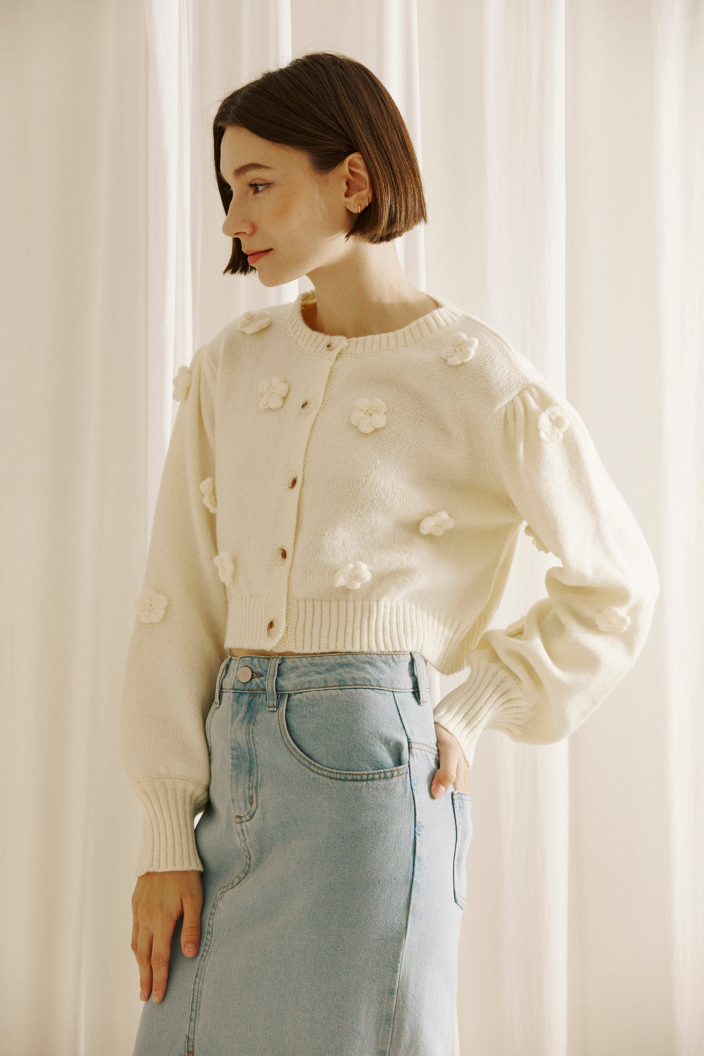 Side view of a woman wearing a cream-colored cardigan with floral embellishments against a white curtain background