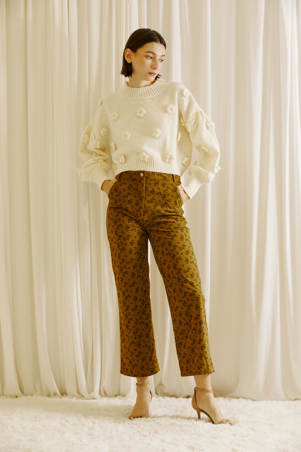 Woman wearing a white sweater with polka dots and patterned brown pants against a white curtain background.