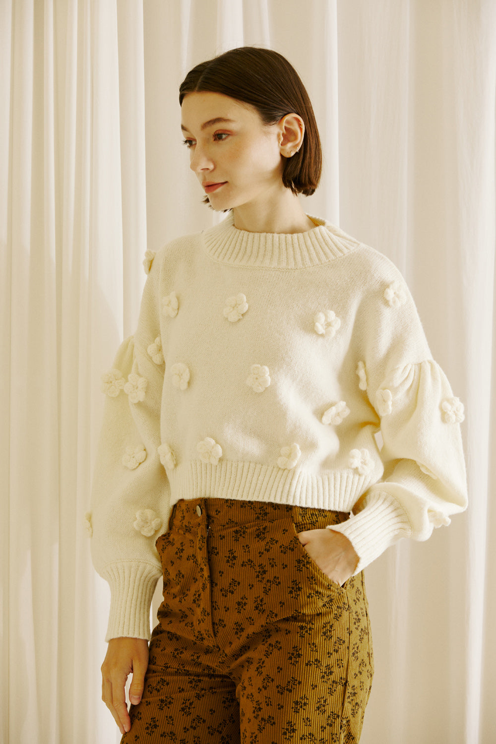 A woman wearing a beige knitted sweater with floral patterns on the sleeves and a V-neckline, paired with brown floral patterned pants.