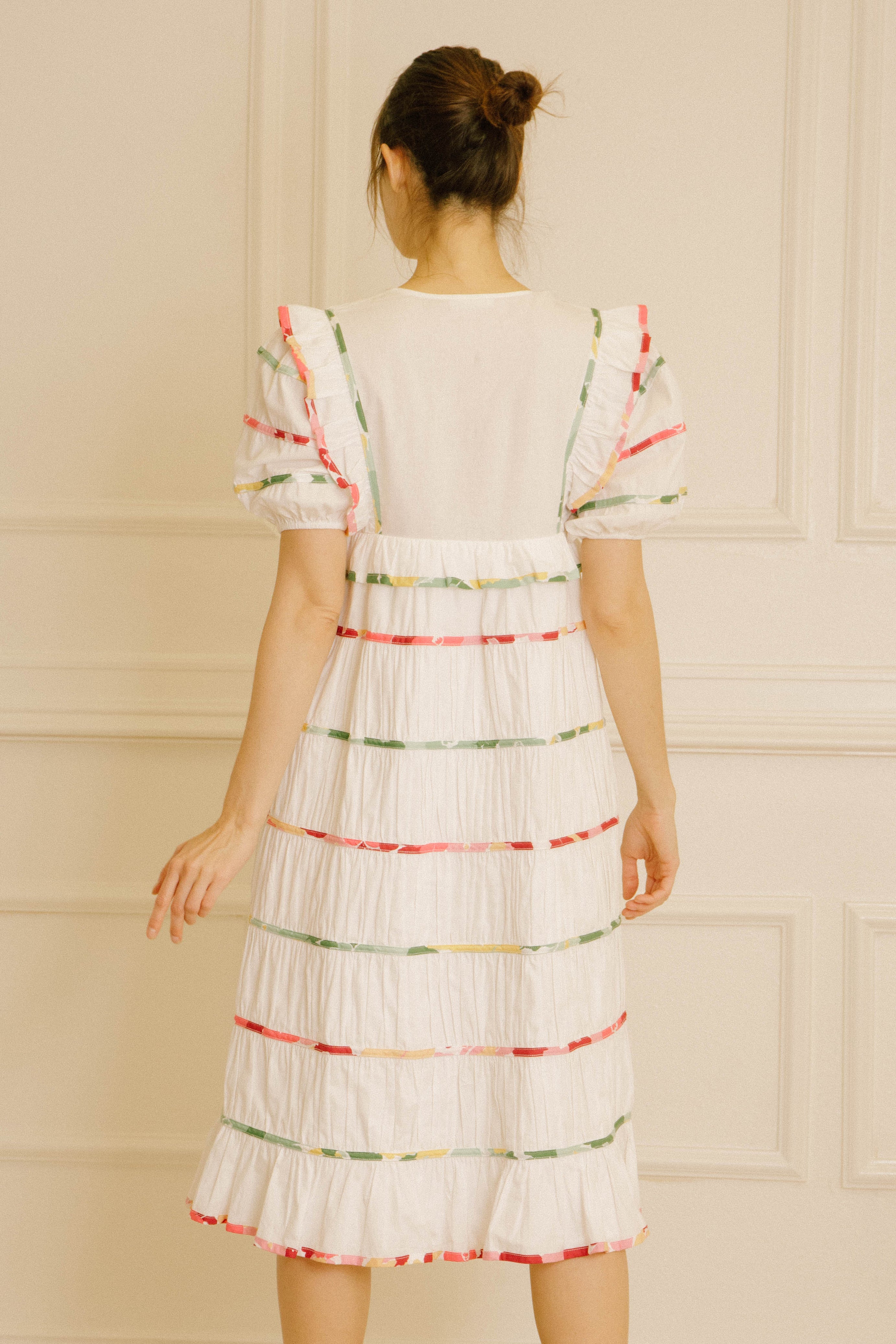 Back view of a woman wearing a white dress with colorful stripes against a beige wall.