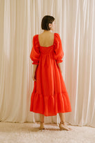 Back view of a woman wearing a red midi flowy dress standing against a white curtain backdrop