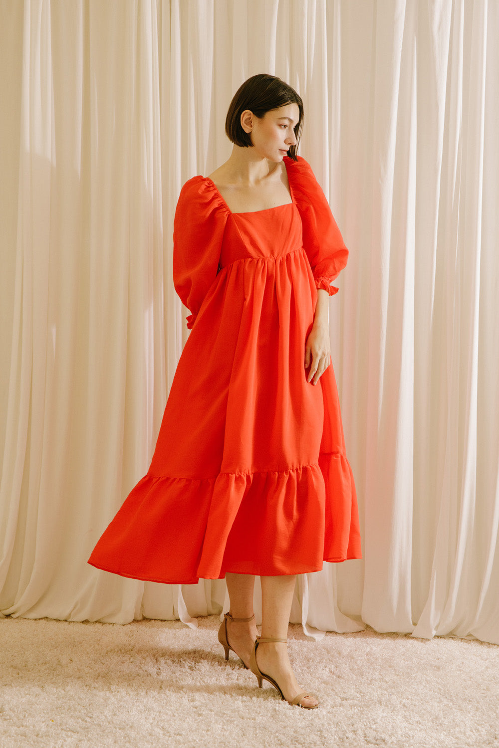 Side view of a woman wearing a red midi flowy dress standing against a white curtain backdrop