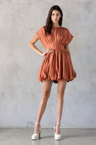 Woman wearing a rust-colored dress against a gray concrete wall.