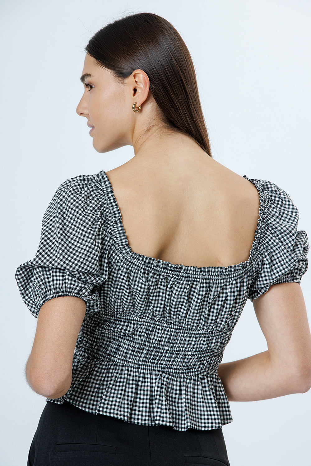 Back view of a woman wearing a black and white checkered blouse with puff sleeves on a plain background
