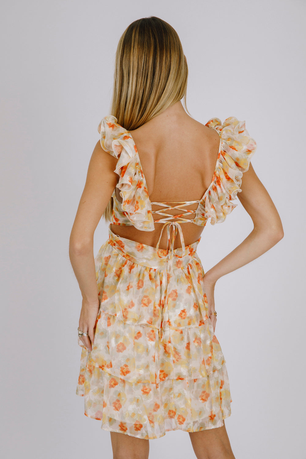 Back view of a woman wearing an orange and cream floral dress with an open back with a crisscross back tie against a plain background