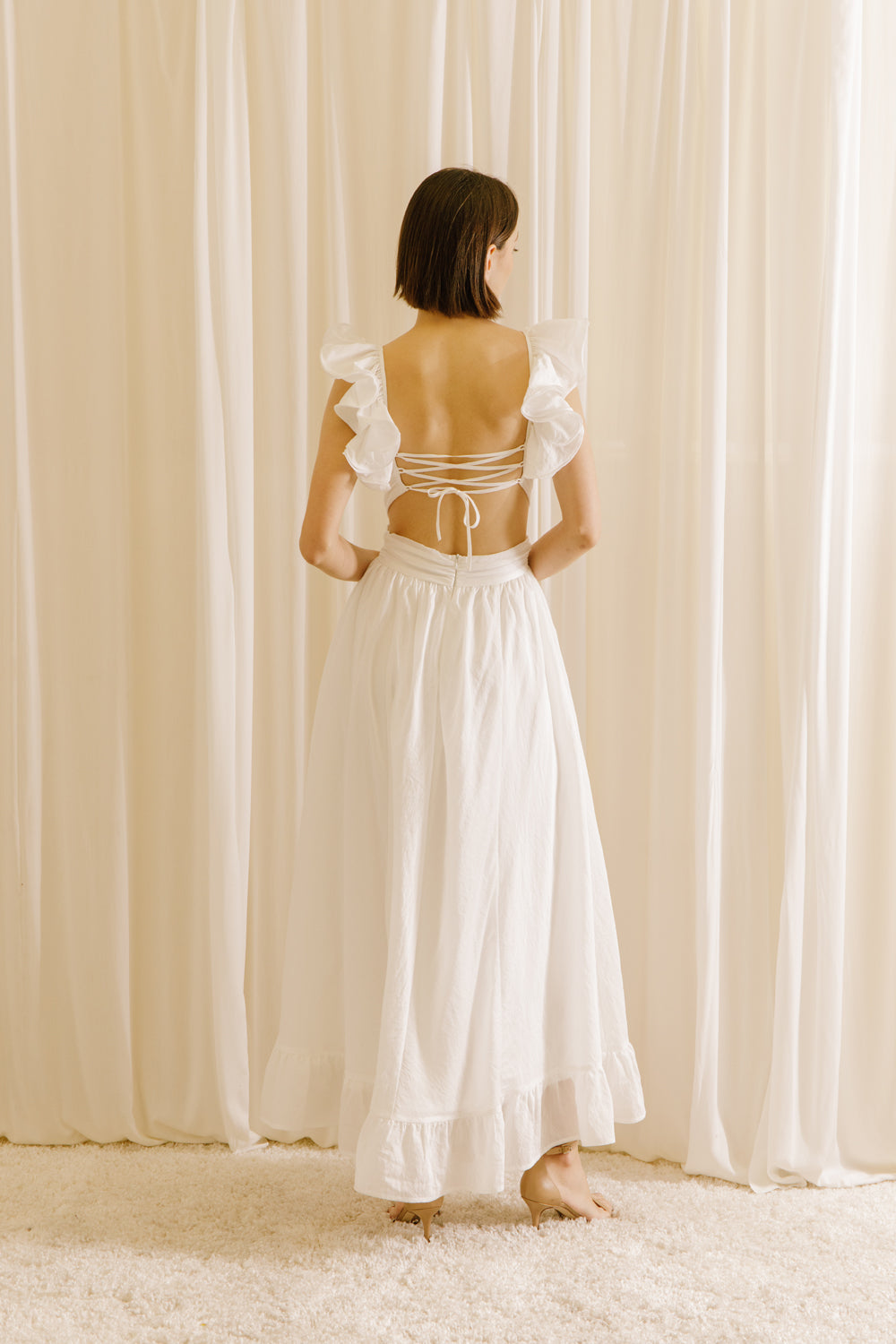 Back view of a woman wearing a white maxi dress with ruffled straps, and an empire waist. The dress features an open back with a criss-cross tie and a back zipper.
