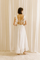 Back view of a woman wearing a white maxi dress with ruffled straps, and an empire waist. The dress features an open back with a criss-cross tie and a back zipper.