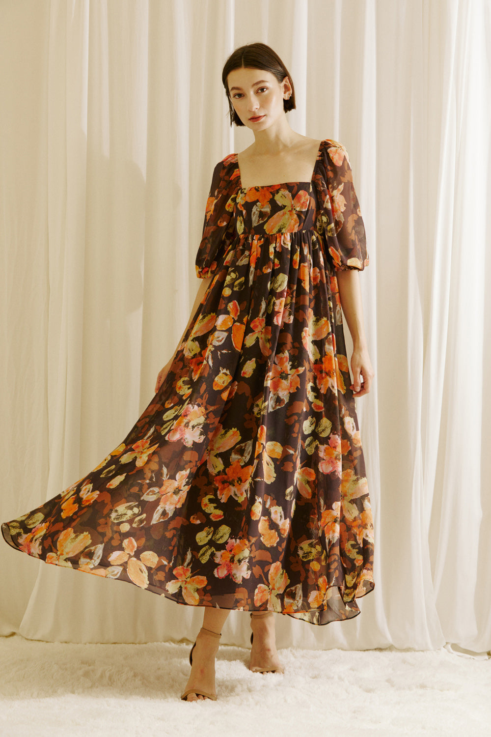 Woman wearing a brown floral dress and beige shoes standing against a white curtain backdrop
