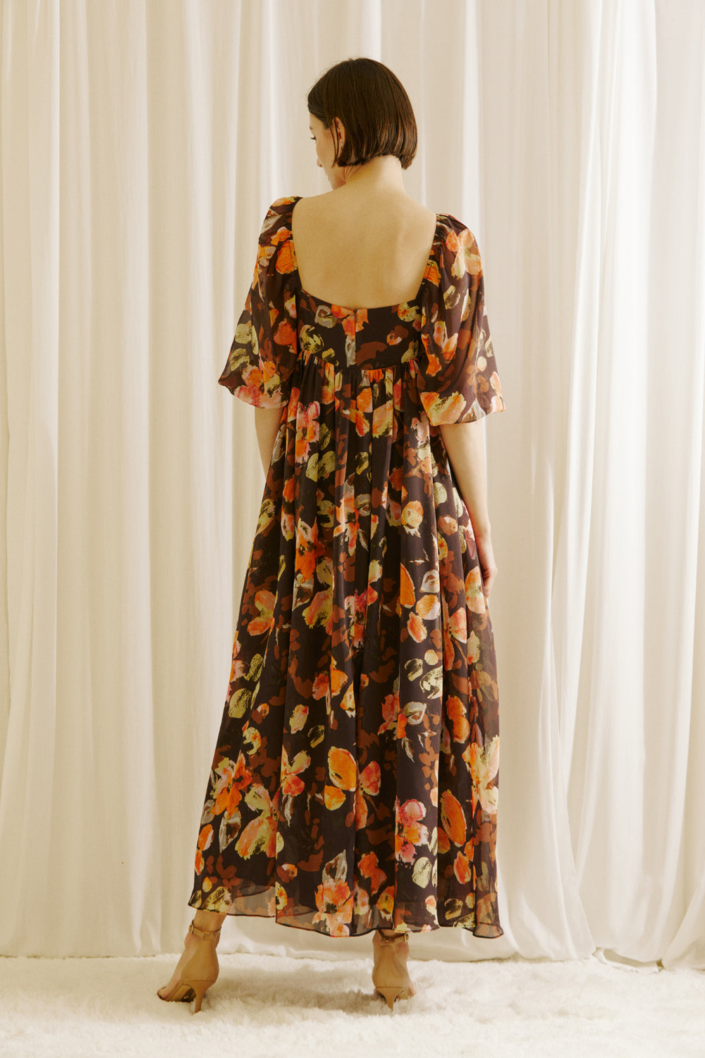 Back view of a woman wearing a brown floral dress standing against a white curtain backdrop