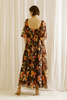 Back view of a woman wearing a brown floral dress standing against a white curtain backdrop