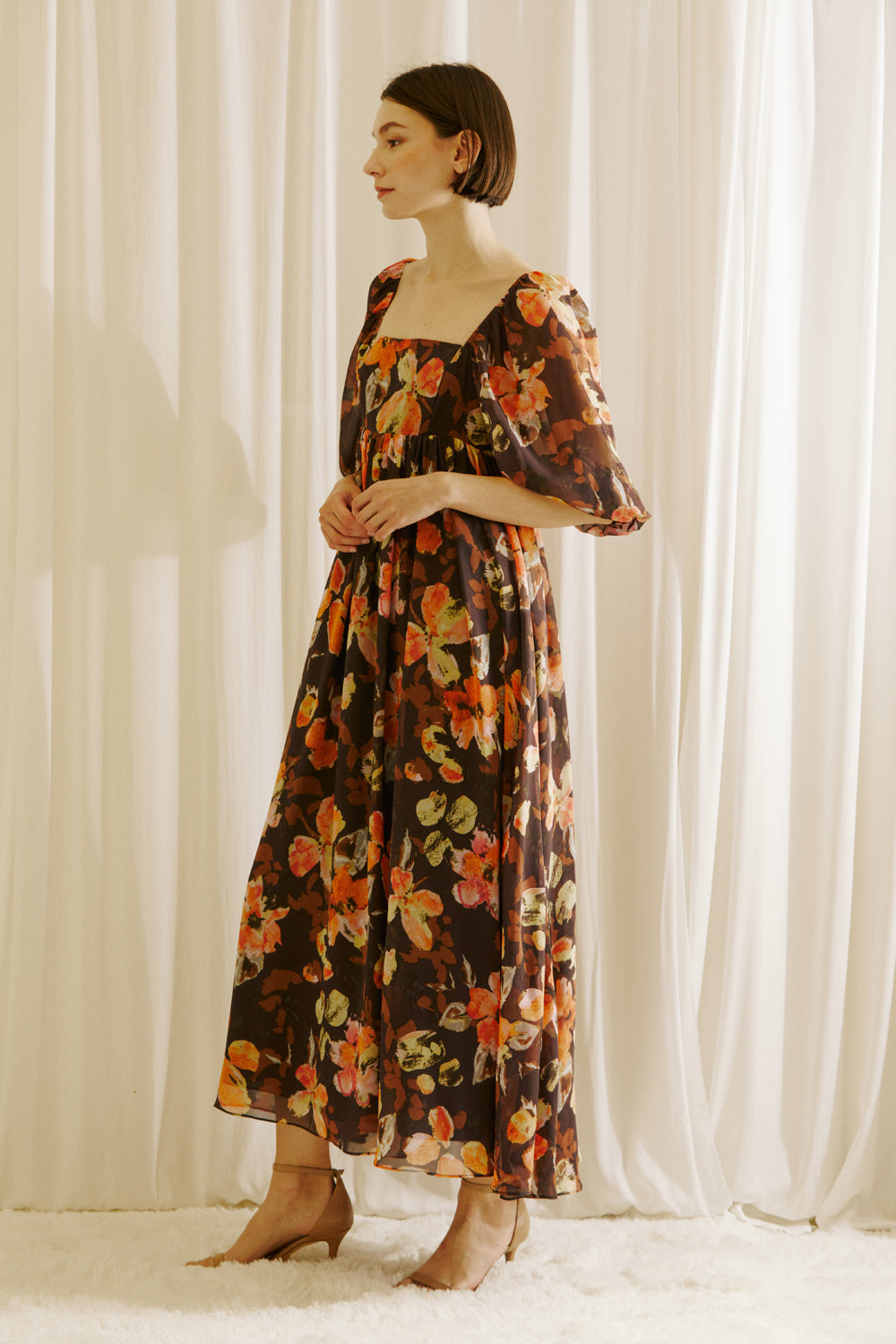 Side view of a woman wearing a brown floral dress standing against a white curtain backdrop