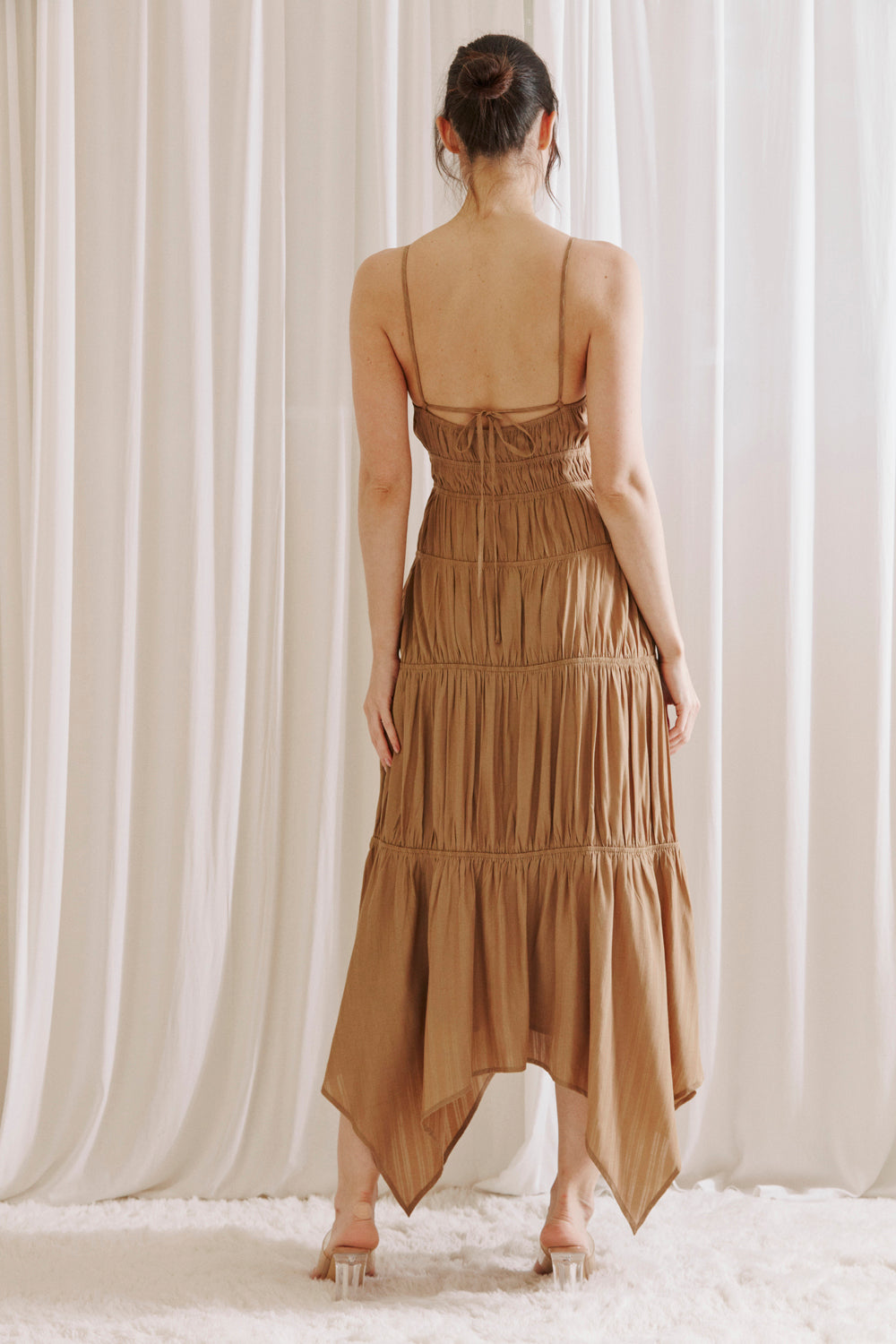 Back view of a woman wearing a brown dress with asymmetrical Hem standing against a white curtain background
