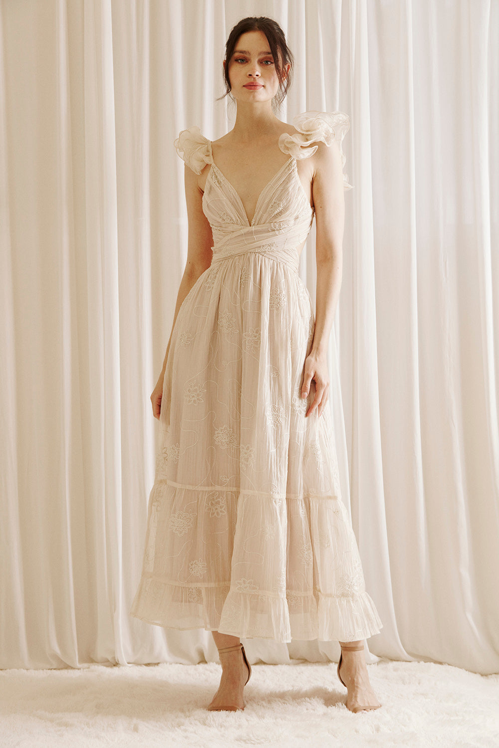 A woman wearing a beige maxi dress with embroidered flowers, featuring a plunging neckline and ruffled straps.